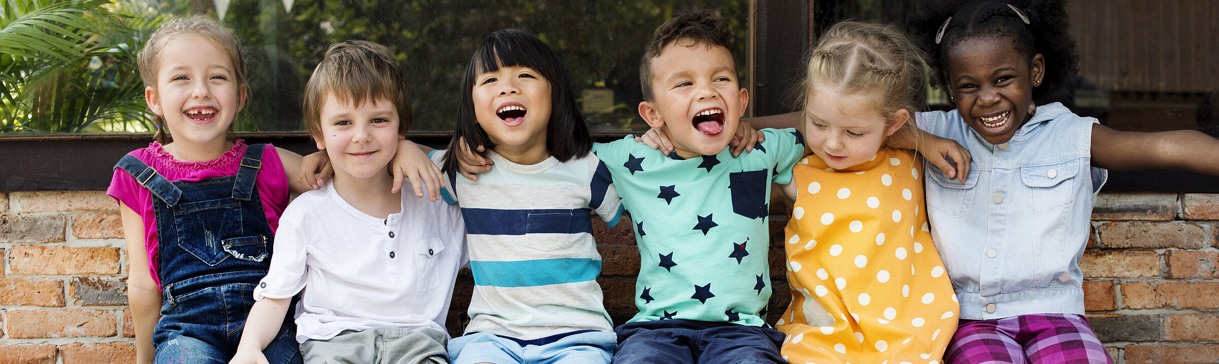 Sechs Kinder sitzen auf einer Bank. Sie haben die Arme um die Schultern voneinander gelegt. Sie haben unterschiedliche Hautfarben und lachen in die Kamera.