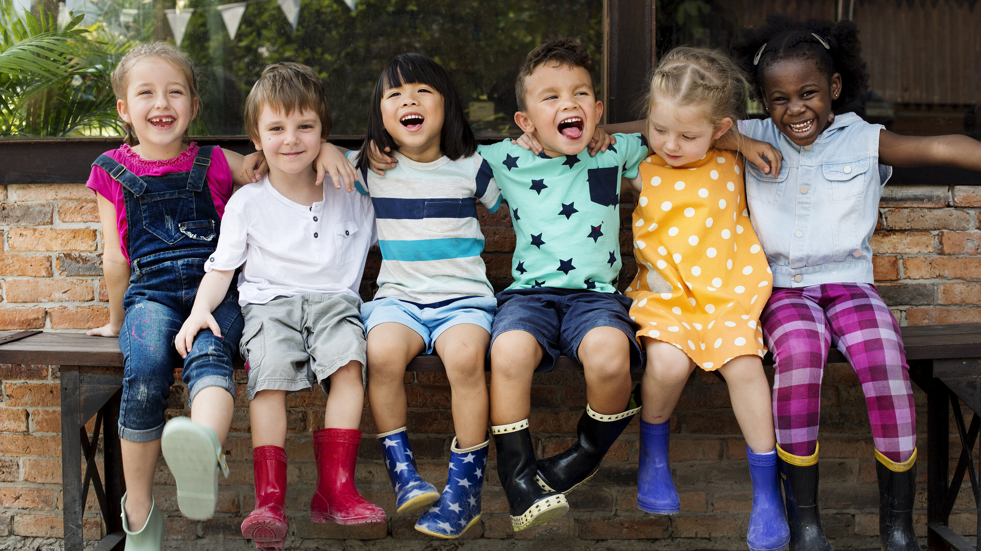 Sechs Kinder sitzen auf einer Bank. Sie haben die Arme um die Schultern voneinander gelegt. Sie haben unterschiedliche Hautfarben und lachen in die Kamera.
