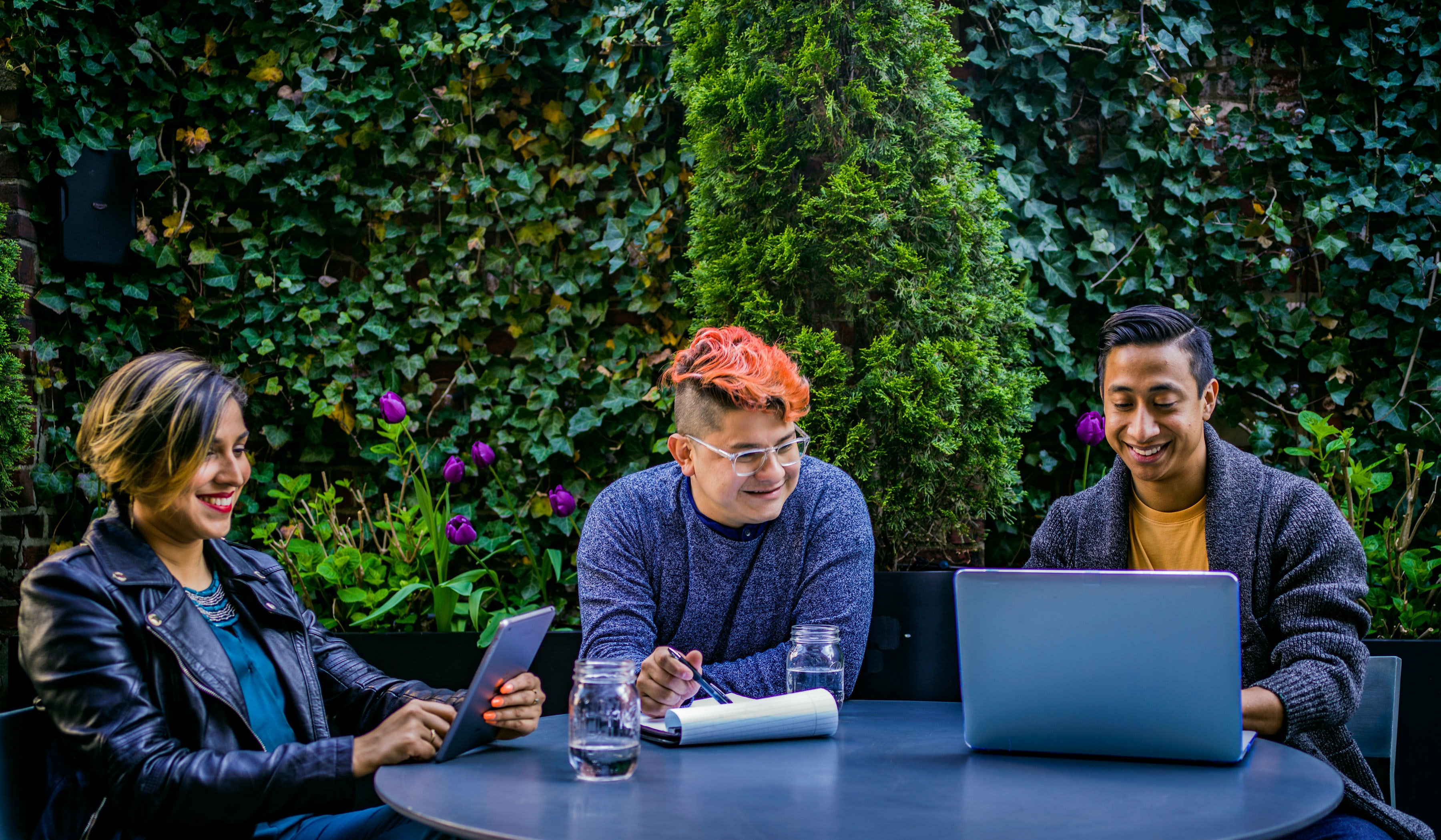 drei Menschen arbeiten draußen an einem Tisch, es steht ein Laptop auf dem Tisch